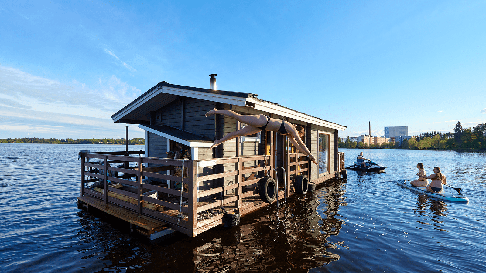 Person jumps to the lake from a sauna raft, two persons are SUP boarding and one person is driving a jet ski.