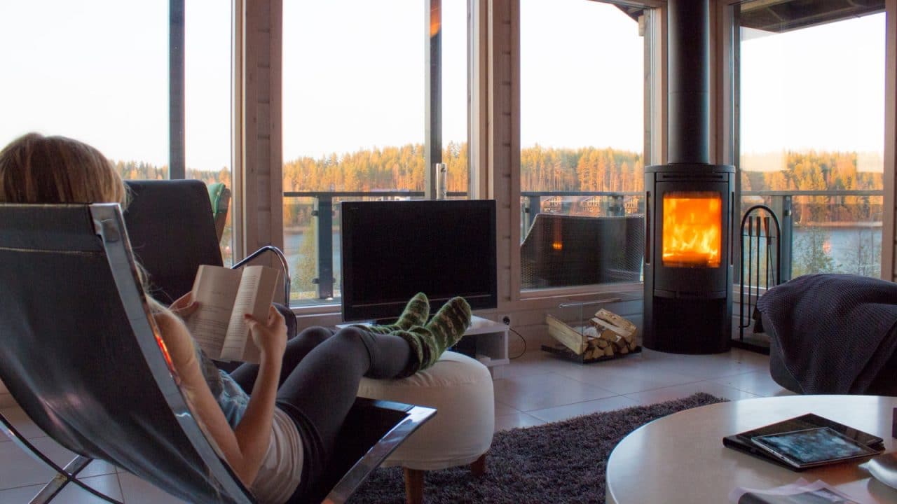 Henkilö istuu mökin nojatuolissa kirjaa lukien. Takassa on tuli ja ikkunoista avautuu maisema järvelle ja metsään.