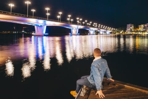 Henkilö istuu laiturilla ja katsoo valaistun Kuokkalan sillan suuntaan.