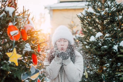 Henkilö seisoo koristeltujen joulukuusien keskellä ja puhaltaa käsissään olevaa lunta.