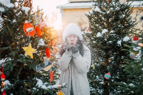 Eine Person steht zwischen den geschmückten Weihnachtsbäumen im alten Hof von Toivola und bläst den Schnee in ihren Händen in die Luft.