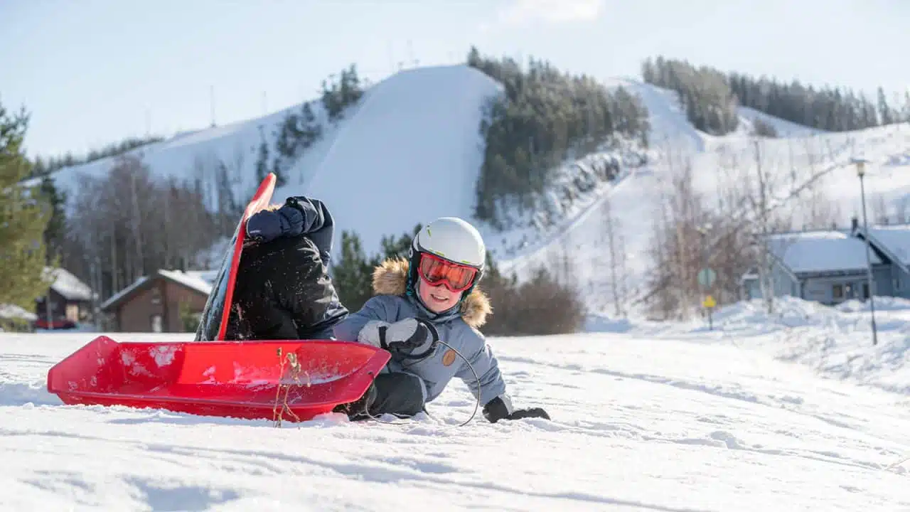 Kaksi lasta pulkan ja liukurin kanssa Himoksella, taustalla Himoksen rinteet ja mökkejä.