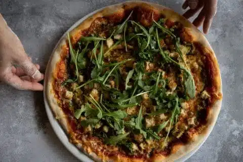 Kaksi kättä pizzajauhoissa laittamassa pizzaa pöydälle.