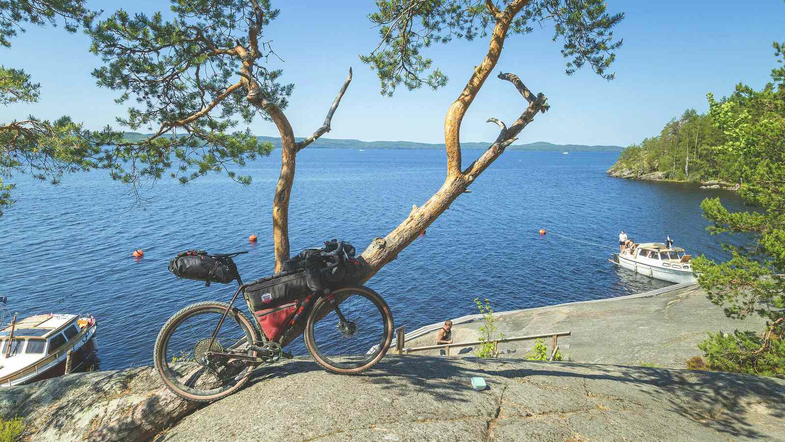 Polkupyörä on rennosti tuettu mäntyä vasten pienen kallion huipulla, ja sieltä avautuu näkymä järvelle, jossa on kaksi venettä kallion alla.