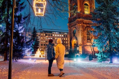 Two persons are standing and holding hands at a snowy Church Park in Jyväskylä. In the background there are some light installations on trees and on the church.