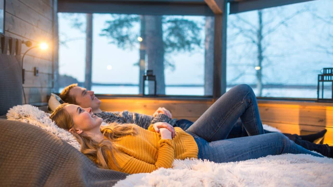 Two people lie on a bed in a glass igloo.