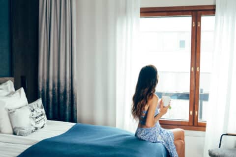 A person is sitting on a bed in a hotel room, coffee mug in hand, looking out of the window.