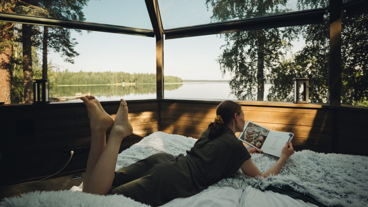 Henkilö lukemassa kirjaa sängyllä lasi-iglussa kesällä.
