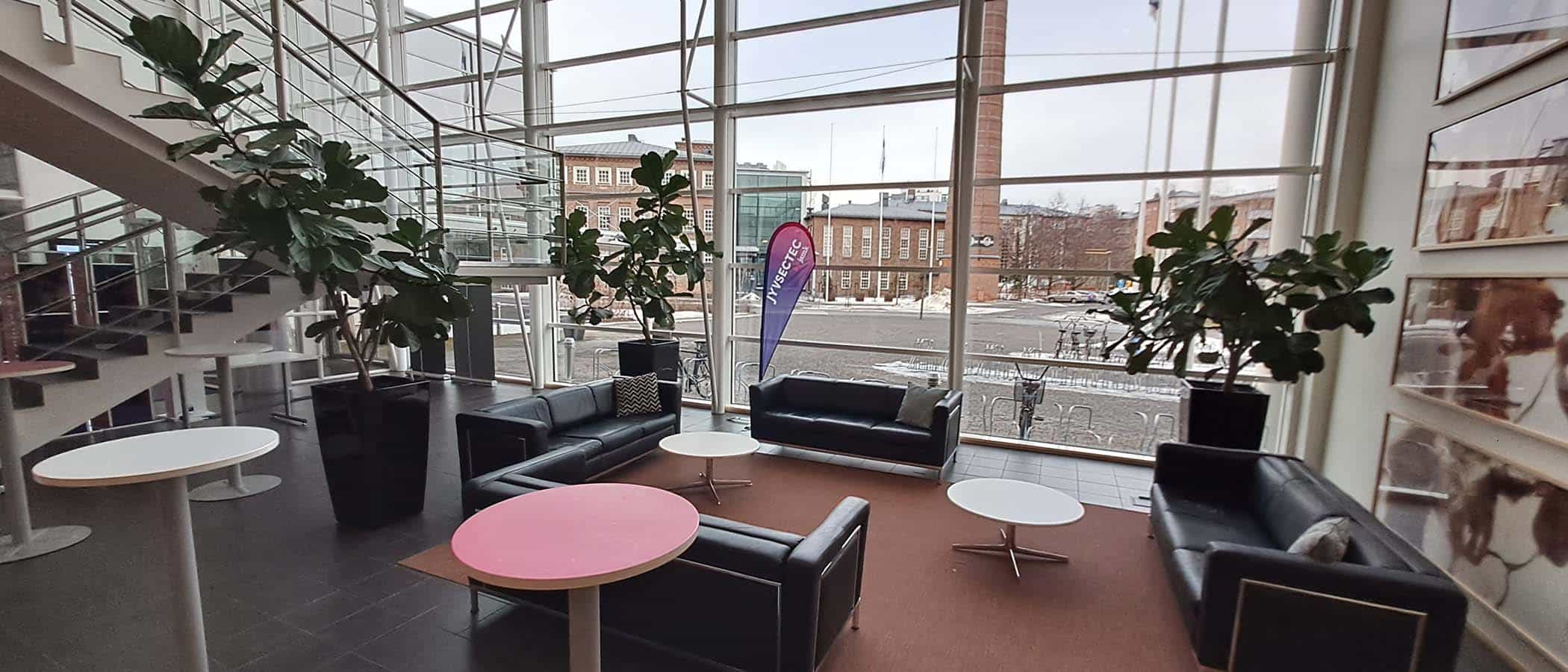 Isot lasi-ikkunat aulassa, mustia nahkasohvia vaalean maton ympärillä, viherkasveja ja pystypöytiä ympärillä, iso portaikko vasemmalla