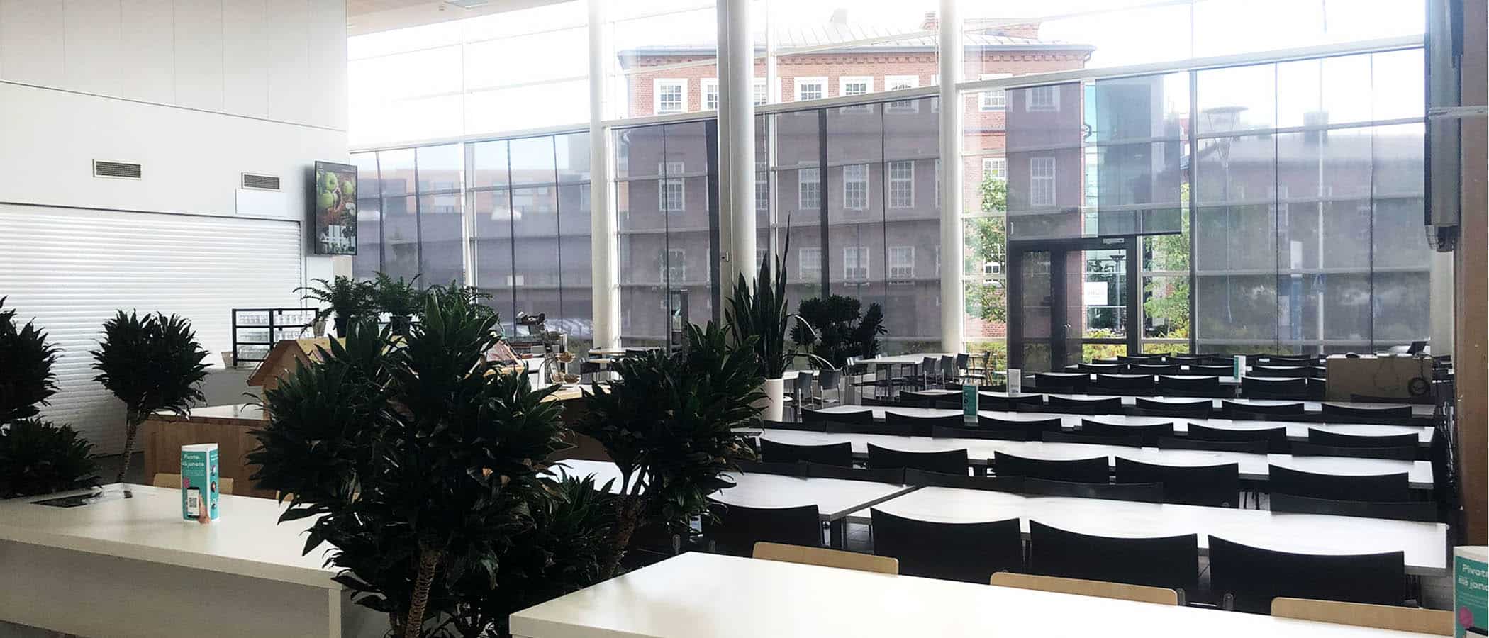 Isot lasi-ikkunat, valkeita pöytiä rivissä, ja niiden ympärillä tuoleja. Muutama viherkasvi