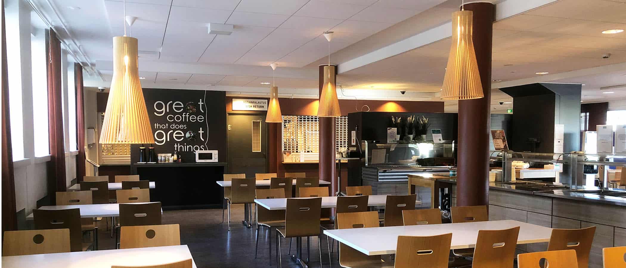 Kahvilassa valkeita neliskanttisia pöytiä  riveissä, ja niiden ympärillä puisia tuoljea. Pöytien yläpuolella riippuvalaisimia. Tarjoilulinjasto näkyy oikealla