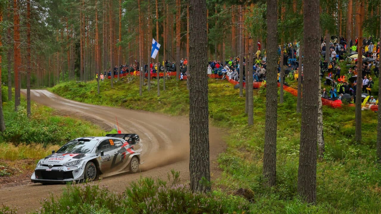 A rally car drives through a pine forest with the crowd cheering in the background.