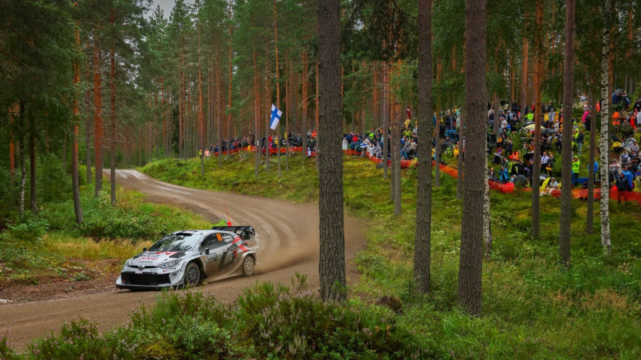 Ralliauto ajaa soratiellä yleisön hurratessa taustalla.