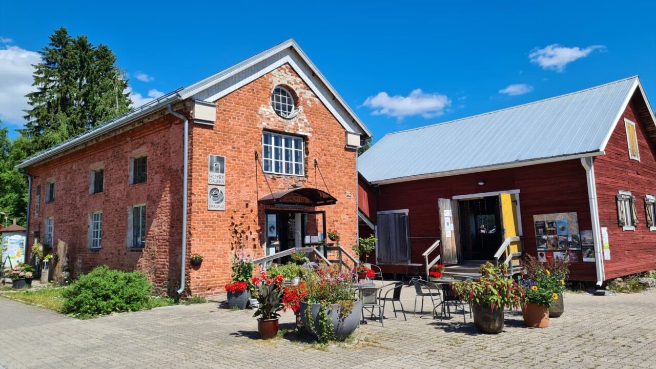 A red brick building with flower plantings and terrace chairs in front of it. Next to it, a red wooden building