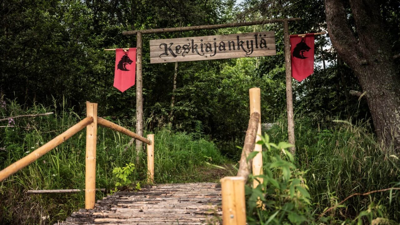 A wooden bridge and gate with a hanging sign saying Keskiajankylä