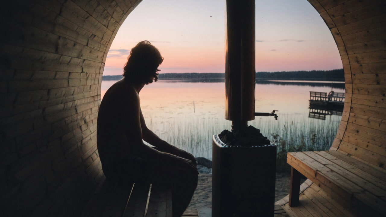 Henkilö istuu tynnyrisaunan lauteilla auringonlaskun maalaaman maiseman edessä.