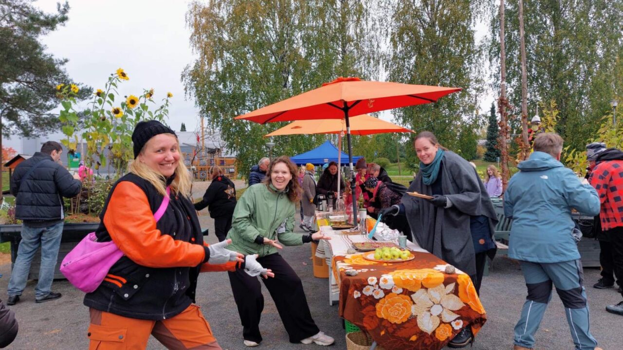 Kuvassa näkyy iloisia henkilöitä esittelemässä pöytää, jossa on omenoita ja leivonnaisia sekä juomia.