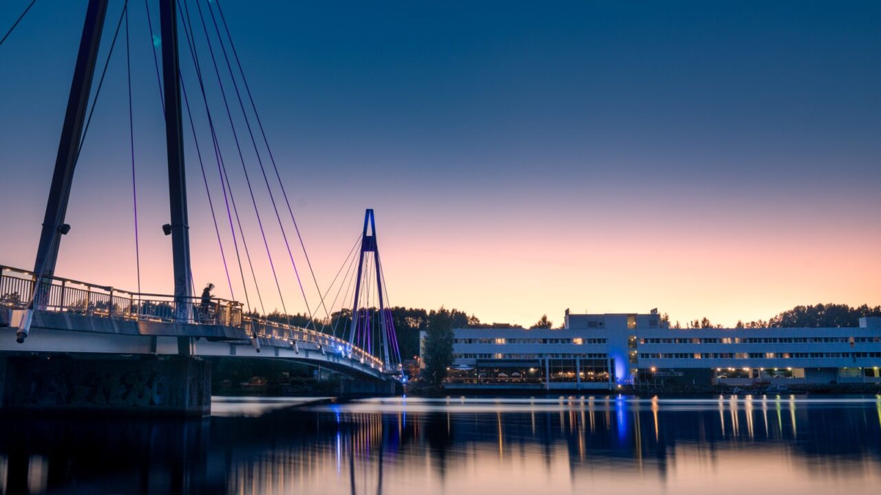 Kuvassa näkyy valaistu silta sekä rakennus ja järveä auringonlaskun aikaan.