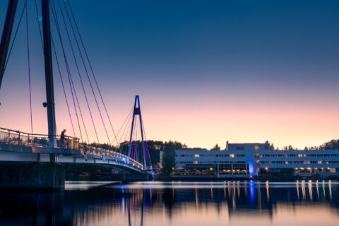 Kuvassa näkyy valaistu silta sekä rakennus ja järveä auringonlaskun aikaan.