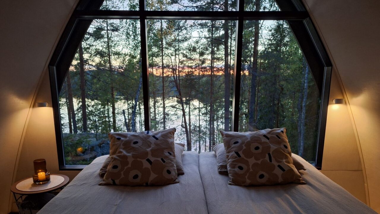 Kaksi sänkyä metsämajoitteessa, jonka suuresta ikkunasta aukeaa metsäinen järvimaisema.